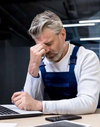 Gestresster Handwerker sitzt am Schreibtisch und wirkt von seinen Unternehmenszahlen und betriebswirtschaftlichen Kennzahlen überfordert.